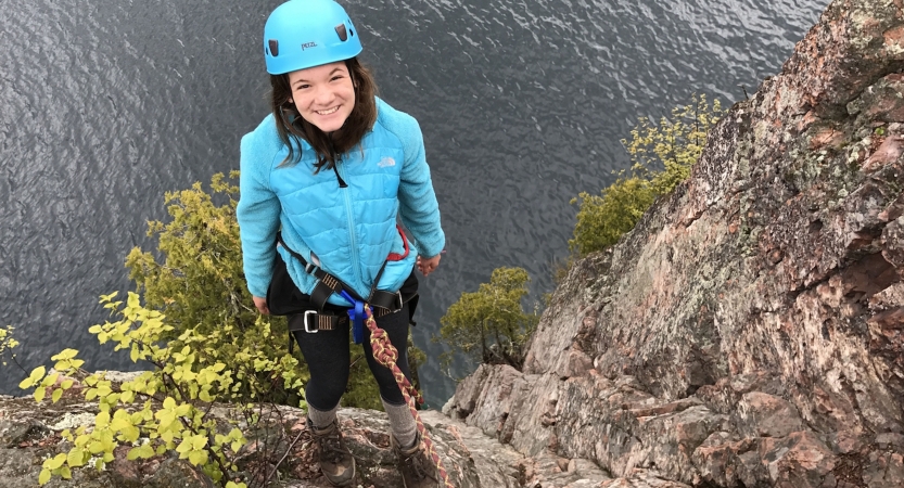 A young person wearing safety gear is secured by ropes as they stand on the edge of a cliff, high above a body of water.